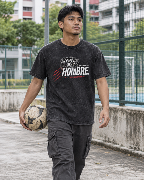 Asian male model wearing the HOMBRE x Balestier Khalsa FC Tiger Instinct regular-fit washed black tee beside a football court